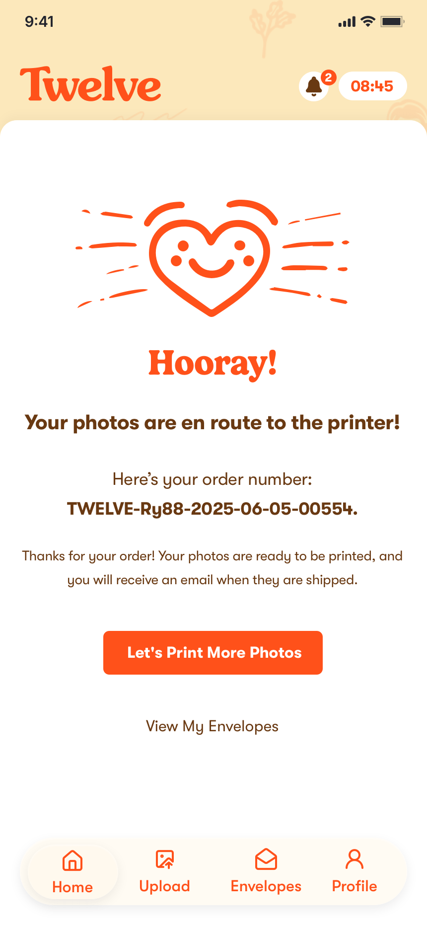 The order-confirmation screen: a hand-drawn smiling heart above the line &ldquo;Hooray! Your photos are en route to the printer!&rdquo;, the order number printed beneath it, a note that an email will follow when the prints ship, and buttons reading &ldquo;Let&rsquo;s Print More Photos&rdquo; and &ldquo;View My Envelopes&rdquo;.