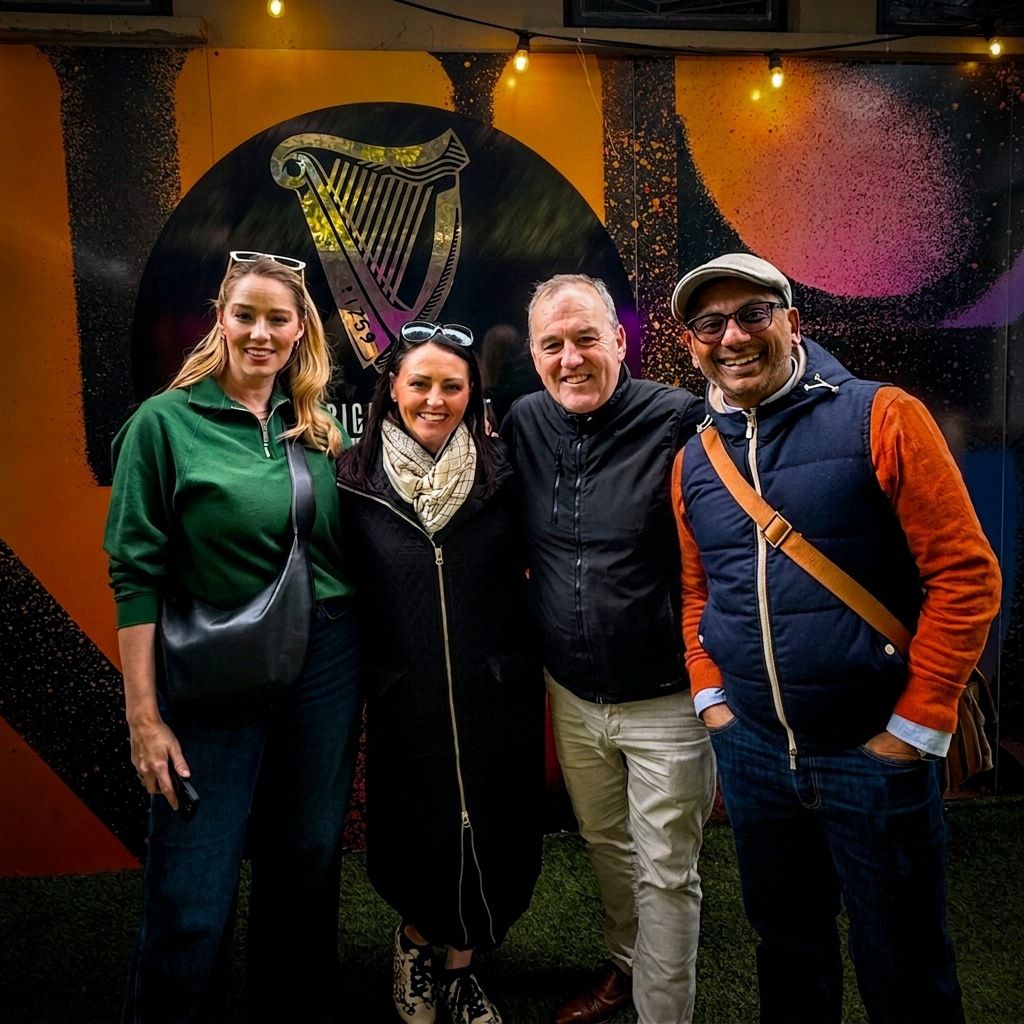 Four people standing shoulder to shoulder, smiling at the camera, in front of a large painted Guinness harp on a hoarding, under string lights on artificial grass.