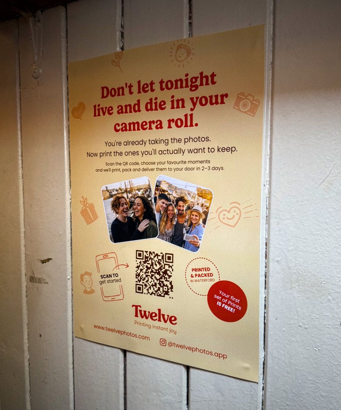 A Twelve Photos poster pasted on a painted wooden wall in low light, headed &ldquo;Don&rsquo;t let tonight live and die in your camera roll.&rdquo; Below it: &ldquo;You&rsquo;re already taking the photos. Now print the ones you&rsquo;ll actually want to keep&rdquo;, two snapshots of friends laughing, a QR code marked &ldquo;scan to get started&rdquo;, and the Twelve wordmark above the line &ldquo;printing instant joy&rdquo;.