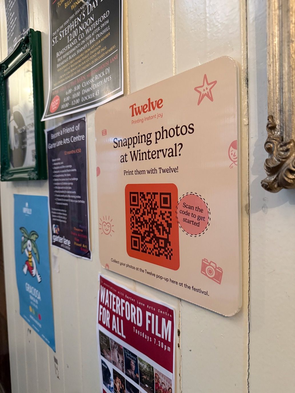 The Twelve QR poster mounted on a painted wooden wall in Waterford among local event flyers, reading &ldquo;Snapping photos at Winterval? Print them with Twelve!&rdquo; above a scannable code.