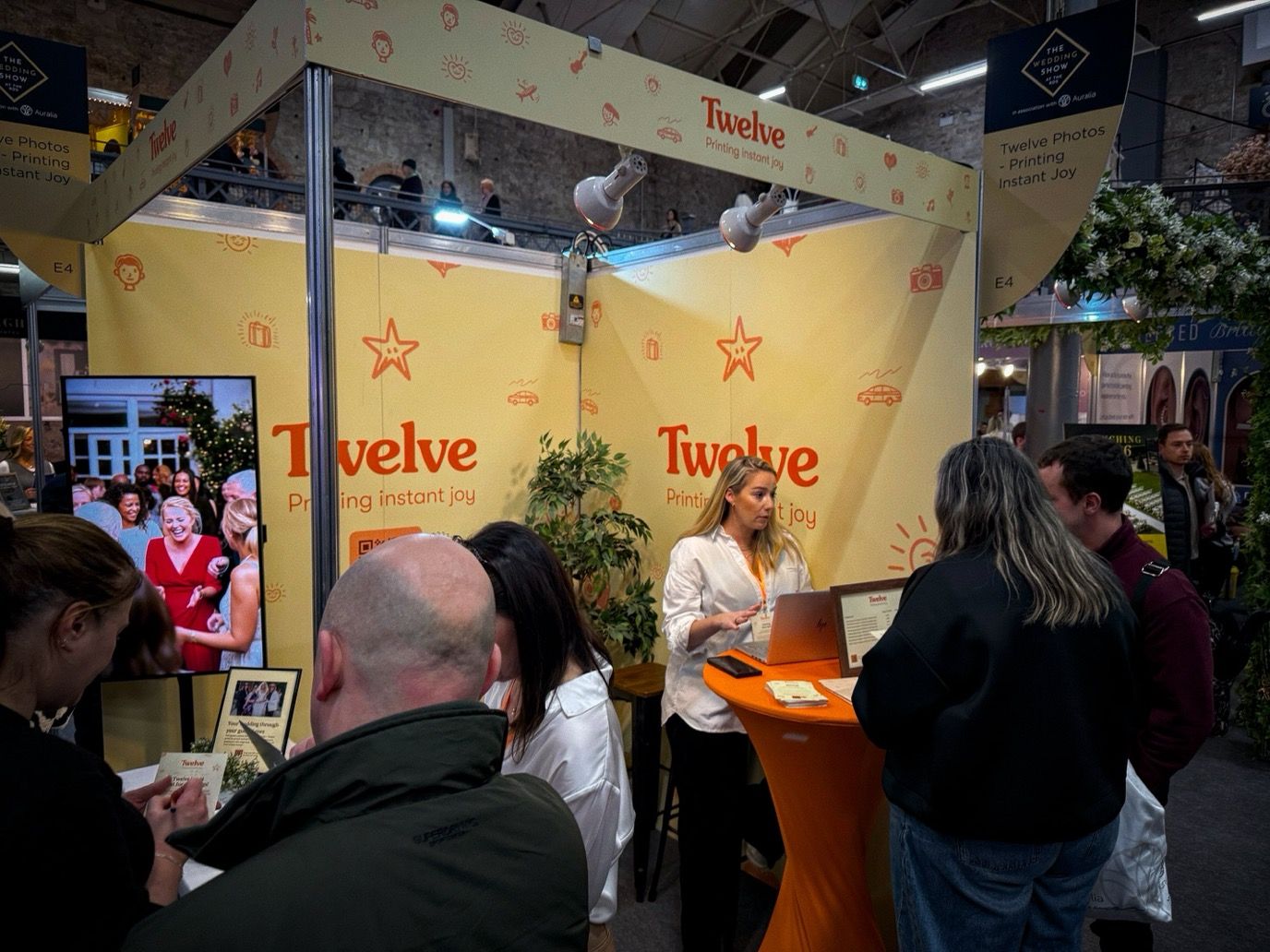 The Twelve Photos stand at The Wedding Show in Dublin: a pale yellow booth printed with the Twelve wordmark and the line &ldquo;Printing instant joy&rdquo;, a screen playing wedding photographs to one side, and visitors gathered at an orange counter talking to a member of the team.