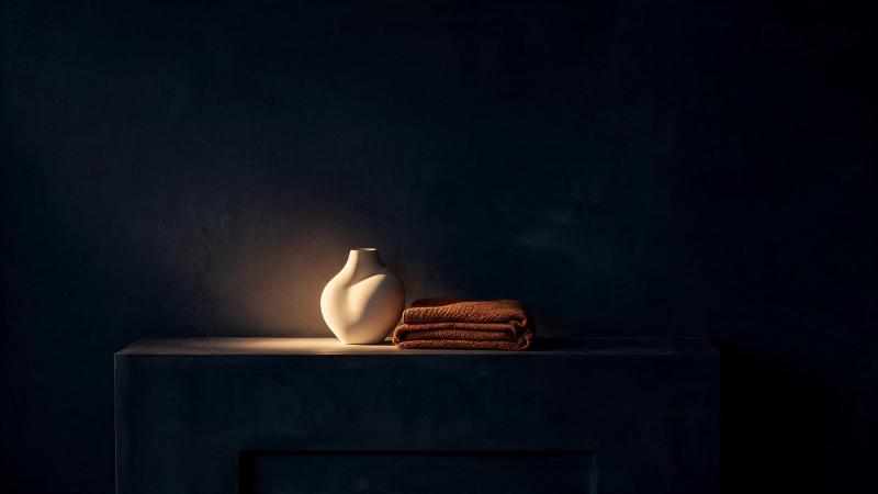 A single ivory vessel and a folded ochre throw lit on a dark console at dusk