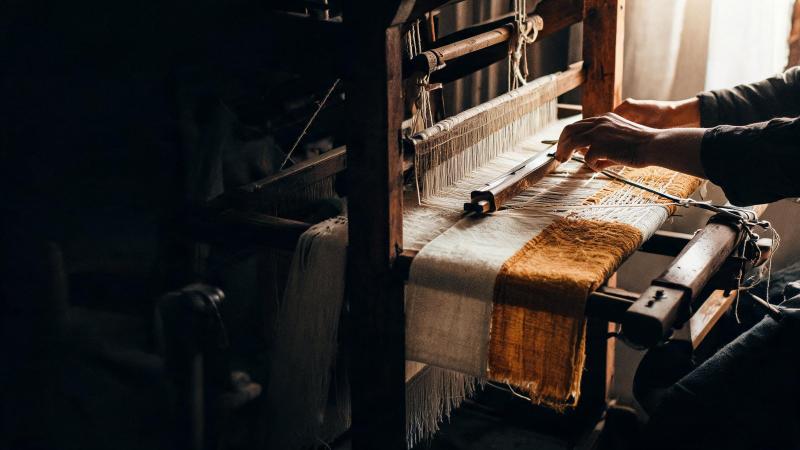 A hand loom with an ochre textile half woven on it, hands at work in warm light