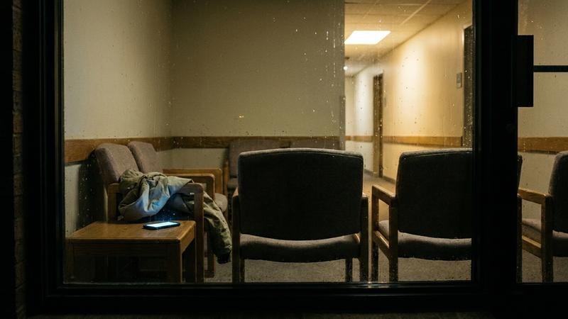 An empty late-night waiting area seen from the street, one corridor light left on