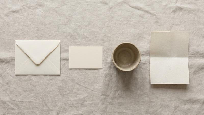 A top-down still life of an enquiry envelope, unmarked appointment card, ceramic cup and face-down follow-up note arranged left to right on pale linen
